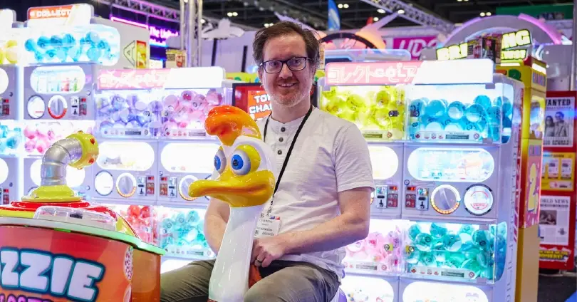 Smiling man with glasses in a white shirt sits on a duck-shaped arcade ride, surrounded by colorful claw machines at a game booth