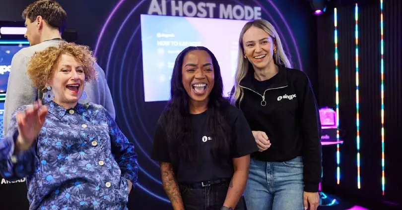 Three people smile and pose at an AI event, with neon lights and a screen reading 'AI HOST MODE' in the background.